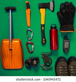 Kit Of Gear For Hiking, Adventure And Survival In Wilderness During Winter - Ax, Avalanche Shovel, Gloves, Gps Receiver And Other Items Lie On A Green Background. Flat Lay, Top View.