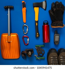 Kit Of Gear For Hiking, Adventure And Survival In Wilderness During Winter - Boots, Avalanche Shovel, Gloves, Gps Receiver, Headlamp And Other Items Lie On A Blue Background. Flat Lay, Top View.