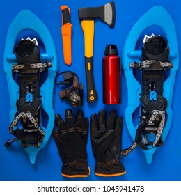 Kit Of Gear For Hiking, Adventure And Survival In Wilderness During Winter - Ax, Snowshoes, Gloves And Other Items Lie On A Blue Background. Flat Lay, Top View.