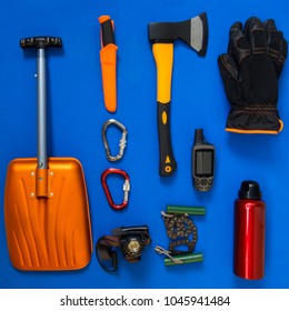 Kit Of Gear For Hiking, Adventure And Survival In Wilderness During Winter - Ax, Avalanche Shovel, Gloves, Gps Receiver And Other Items Lie On A Blue Background. Flat Lay, Top View.