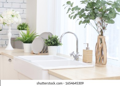 kitchen area with artificial flowers in flower pots, plates on a wooden stand, a large sink with a tap.