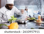 In a kitchen, black chef adding fresh herbs to complete a gourmet dish, balancing flavors with rosemary and other condiments. Kitchen counter stocked with vegetables and food prep essentials.