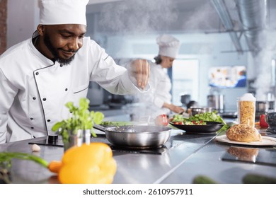 In a kitchen, black chef adding fresh herbs to complete a gourmet dish, balancing flavors with rosemary and other condiments. Kitchen counter stocked with vegetables and food prep essentials.