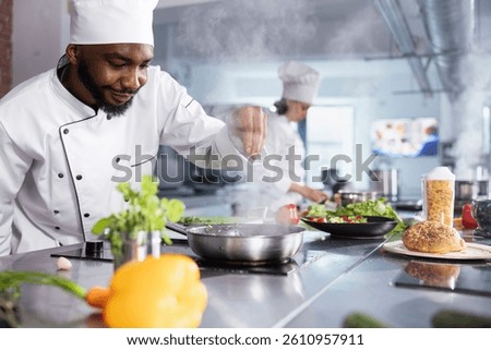 In a kitchen, black chef adding fresh herbs to complete a gourmet dish, balancing flavors with rosemary and other condiments. Kitchen counter stocked with vegetables and food prep essentials.