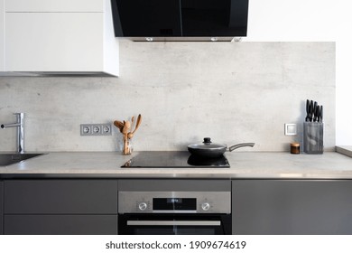 Kitchen With Built In Appliance, Electric Cooker Hood, Stove, Oven And Sink. Frying Pan On Glass Ceramic Surface At Marble Countertop Close To Wooden Spoons And Knives, Under White Cupboards