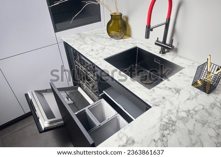 Kitchen with built-in opened dishwasher and pull out drawer for waste bin separate waste collection. Water faucet sink built in compact high pressure laminate HPL countertop undermount installation.