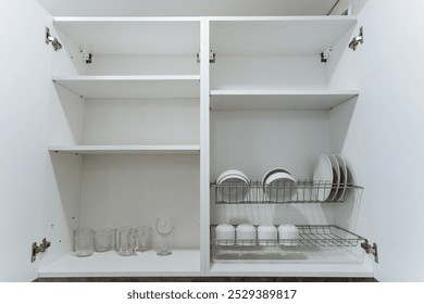 The kitchen cabinet is filled with various tableware items including plates, cups, glasses, and also features a dish rack for drying. It serves a practical purpose in the kitchen space