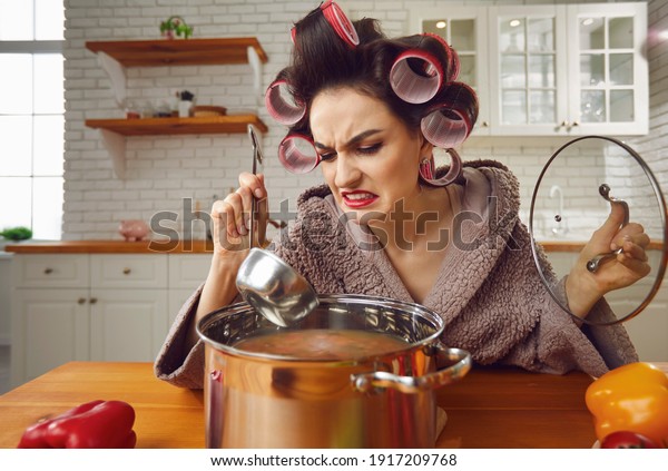 Kitchen calamity. Bad unskilled novice housewife\
cooking food for the first time. Funny woman in hair curlers\
holding ladle and tasting smelly stinky disgusting yucky disastrous\
awful uneatable soup