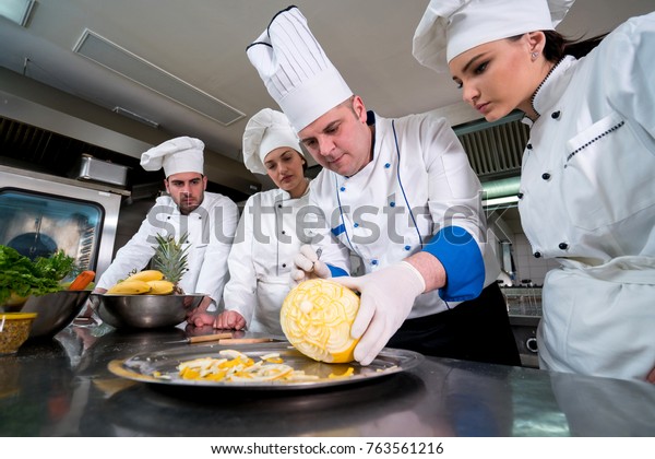 Kitchen chef with young apprentices, teaching to\
make decorative fruit\
basket