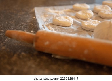 A kitchen countertop with a wooden rolling pin and a sheet of parchment paper. There are round raw biscuits cut and shaped on the tray for baking. The counter is dark brown with black specks.  - Powered by Shutterstock