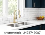 A kitchen detail with a gold faucet in front of a window, white subway tile backsplash, a bowl of fruit on the white marble countertop, and blue cabinets.