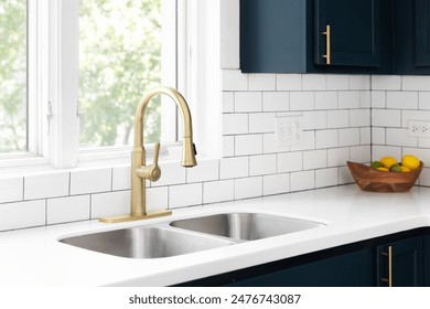 A kitchen detail with a gold faucet in front of a window, white subway tile backsplash, a bowl of fruit on the white marble countertop, and blue cabinets.