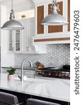 A kitchen detail with metal lights hanging above a large marble island, stainless faucet on a white farmhouse sink, and a marble hexagon tiled backsplash with white cabinets.