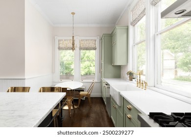 A kitchen detail with sage green cabinets, white marble countertops, gold faucet on an apron sink, and gold chandelier hanging above a dining table.