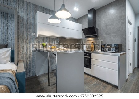 A kitchen and dining area are shown in a small apartment. The space includes a countertop, stove, and dining items. Natural light enters through the windows