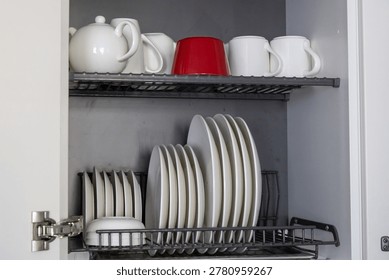 A kitchen dish rack stores an assortment of white ceramic plates and bowls on the bottom shelf, while the top shelf holds several white mugs, a teapot, and a single vibrant red cup for a pop of color inside the grey cabinet.