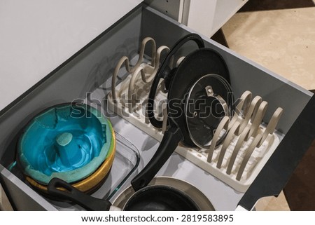 A kitchen drawer containing various cookware items. It includes a blue mixing bowl, several frying pans, and a pot organizer..