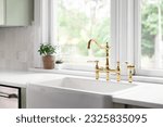 A kitchen faucet in a cozy green kitchen with a gold bridge faucet, white apron sink, tiled backsplash, and white marble countertop.