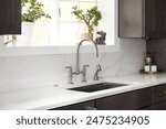 A kitchen faucet detail in a cozy kitchen with a stainless faucet, white marble countertop and backsplash, a bright window, and dark wood cabinets.