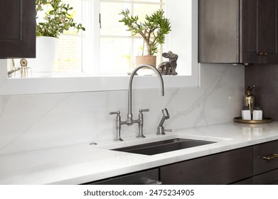 A kitchen faucet detail in a cozy kitchen with a stainless faucet, white marble countertop and backsplash, a bright window, and dark wood cabinets. - Powered by Shutterstock