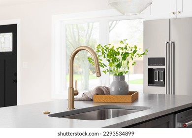 A kitchen faucet detail with a grey stone countertop island, gold faucet, stainless steel appliances, and white cabinets. - Powered by Shutterstock