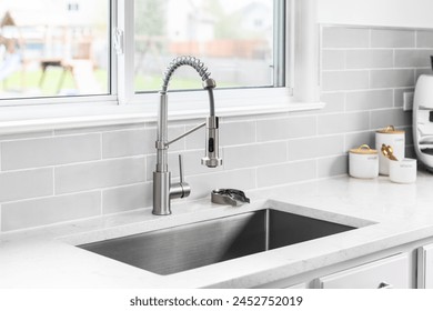 A kitchen faucet detail with a stainless faucet and sink, grey subway tile backsplash, and decorations on the marble countertop.