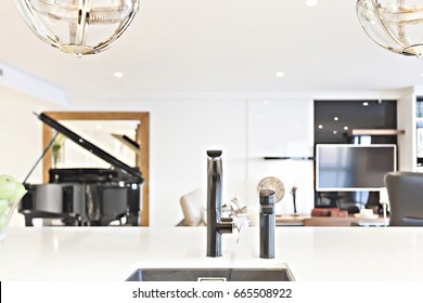 Kitchen Faucet Focusing On The White Counter Top With Blurred Background With White Walls, A Piano And A Television Can Be Seen Blurred From The Behind