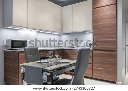 A kitchen features a dining table with plates and glasses ready for a meal. Cabinets and appliances are arranged in a sleek design. Soft lighting adds to the setting
