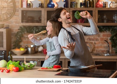 Kitchen Fun. Cheerful Dad And His Little Daughter Singing While Cooking Together, Using Spatula And Broccoli As Microphones