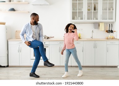 Kitchen Fun. Full Length Of Joyful Black Dad And His Little Daughter Singing And Dancing While Cooking Together At Kitchen, Girl Using Whisk As Microphone, Father Playing On Leg As On Guitar