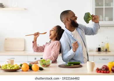 Kitchen Fun. Portrait of positive African American dad and his little daughter singing and dancing while cooking together at the kitchen, using spatula and broccoli as microphones, preparing meal