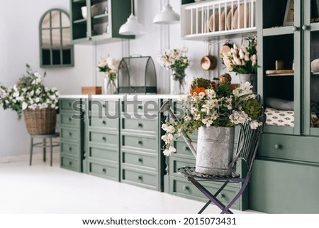 Kitchen with green and white theme, old metal watering pot full of greenery on folding chair, flowers all over counter in blurred background, vintage monochrome furniture