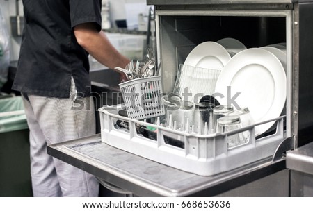 Kitchen hand with an open dishwasher filled with clean white plates in a restaurant kitchen in a catering and hygiene concept