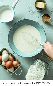 Kitchen - Hand Stirring Pancake Batter In A Bowl. Recipe Ingredients Around (eggs, Milk, Flour, Butter) On Pastel Blue Stone Background. Captured From Above (top View, Flat Lay).