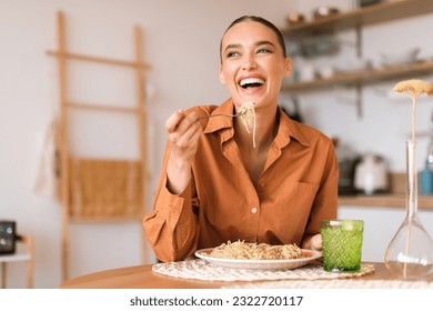 Kitchen happenings. Excited woman eating tasty homemade pasta, enjoying delicious lunch and laughing, sitting in cozy kitchen interior, free space