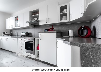 Kitchen At Home In Classic Style, Decorated In White With Marble Fittings And  With Numerous Accessories And Appliances In Stainless Steel Or In Dark Red Color. Floor Is Lined With White Square Tiles.