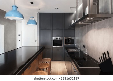 kitchen island with black countertop, bar stools, modern cabinet, induction stove, electric oven. sink with faucet and blue ceiling lamps in studio flat - Powered by Shutterstock