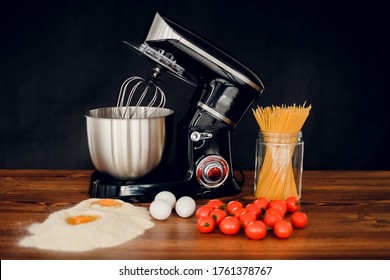 Kitchen Mixer Standing On Wooden Countertop With Separated Eggs, Pasta And Cherry Tomatoes On Dark Background