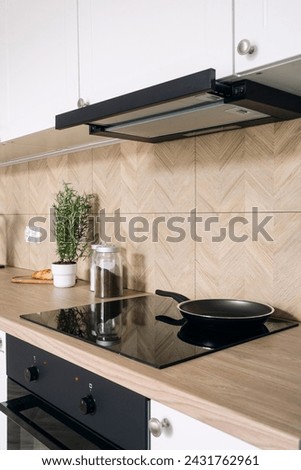 Kitchen with modern interior and household appliances, wooden countertop, integrated glass ceramic electrical hob and cooker hood. Vertical shot of frying pan on induction hob. Cooking food at home 商業照片 © 