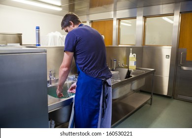 Kitchen Porter Washing Up At Sink In Professional Kitchen