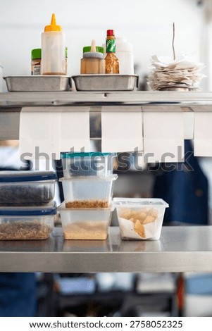 A kitchen prep station with various condiments, sauces, and containers filled with ingredients. A stack of paper plates is visible on a metal shelf.