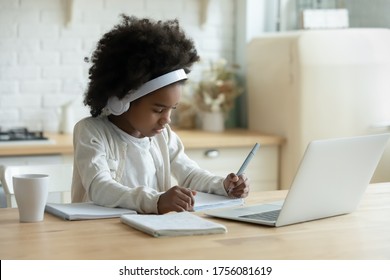 In kitchen schoolgirl do homework, focused little African girl wear headphones watch video lesson using laptop app, interested in on-line web virtual class studying from at home, homeschooling concept
