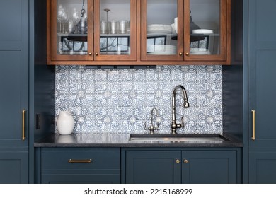 A Kitchen Sink With A Beautiful Pattern Tiled Backsplash With A Chrome Faucet, Black Granite Countertops, And Surrounded By Blue And Wood Cabinets.