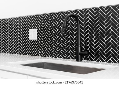 A Kitchen Sink Detail Shot With A Black Faucet, Black Marble Herringbone Backsplash, And A White Granite Countertop.