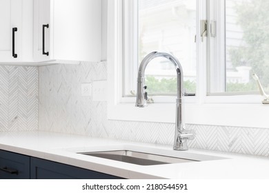 A Kitchen Sink Detail Shot With Blue And White Cabinets, Herringbone Tile Backsplash, And A Chrome Faucet In Front Of A Window.