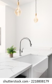 A kitchen sink detail shot with glass pendant lights hanging above the white granite countertop, apron sink, and chrome faucet. 