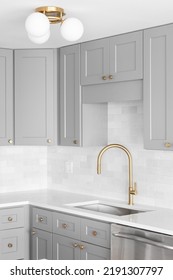 A Kitchen Sink Detail Shot With A Gold Faucet, Marble Backsplash, Grey Cabinets, And Gold Hardware And Light Fixture.