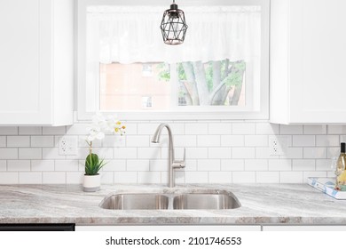 Kitchen Sink Detail Shot With A Marble Countertop, White Cabinets, Stainless Steel Faucet And Sink, And A Light Hanging In Front Of The Window.