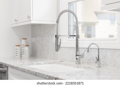 A Kitchen Sink Detail Shot In A Renovated Kitchen With White Cabinets, Chrome Faucet, Marble Herringbone Backsplash, And Granite Countertops.