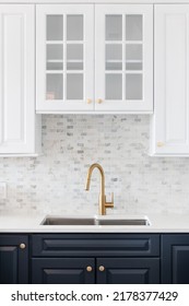 A Kitchen Sink Detail Shot In A White And Blue Kitchen With A Gold Faucet, Marble Countertop, And Small Marble Subway Tile Backsplash.